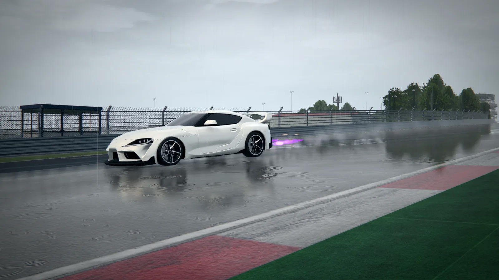A white sports car drifting on a wet circuit beside a red and white kerb and green run-off, with a fence and a pit building behind