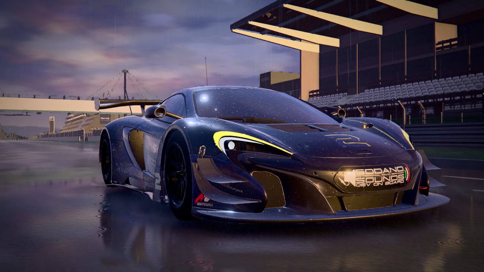 A dark blue GT3 race car with yellow headlight accents on a wet circuit in the evening rain, in front of a grandstand