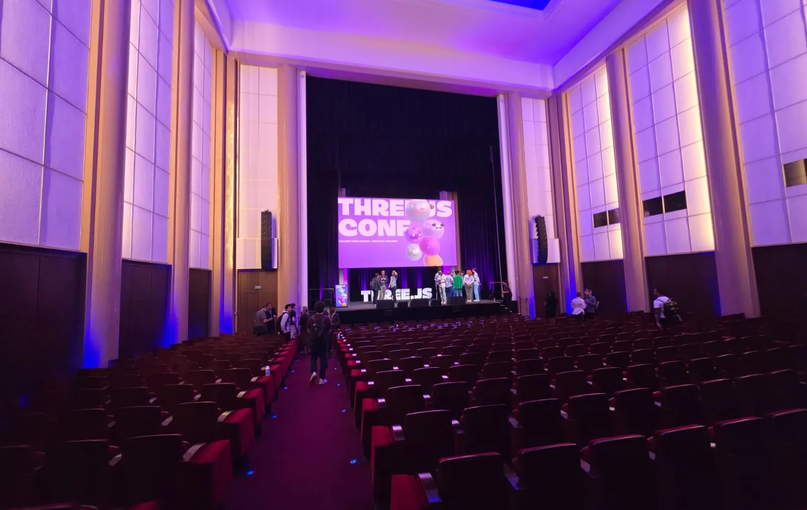 The main hall of the Three.js Conf in Paris, with rows of red seats facing a stage and a purple screen showing the conference logo
